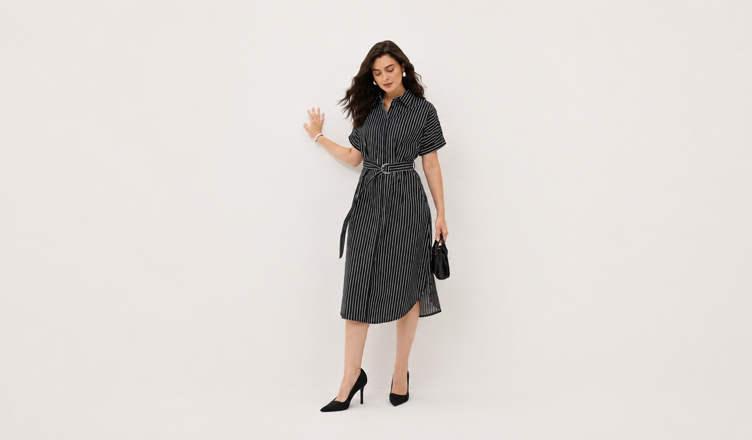 A woman modeling a Salt attire's short-sleeve black and white vertically striped midi shirt dress with a matching tie belt, styled with black heels and a small black handbag against a plain white studio background.